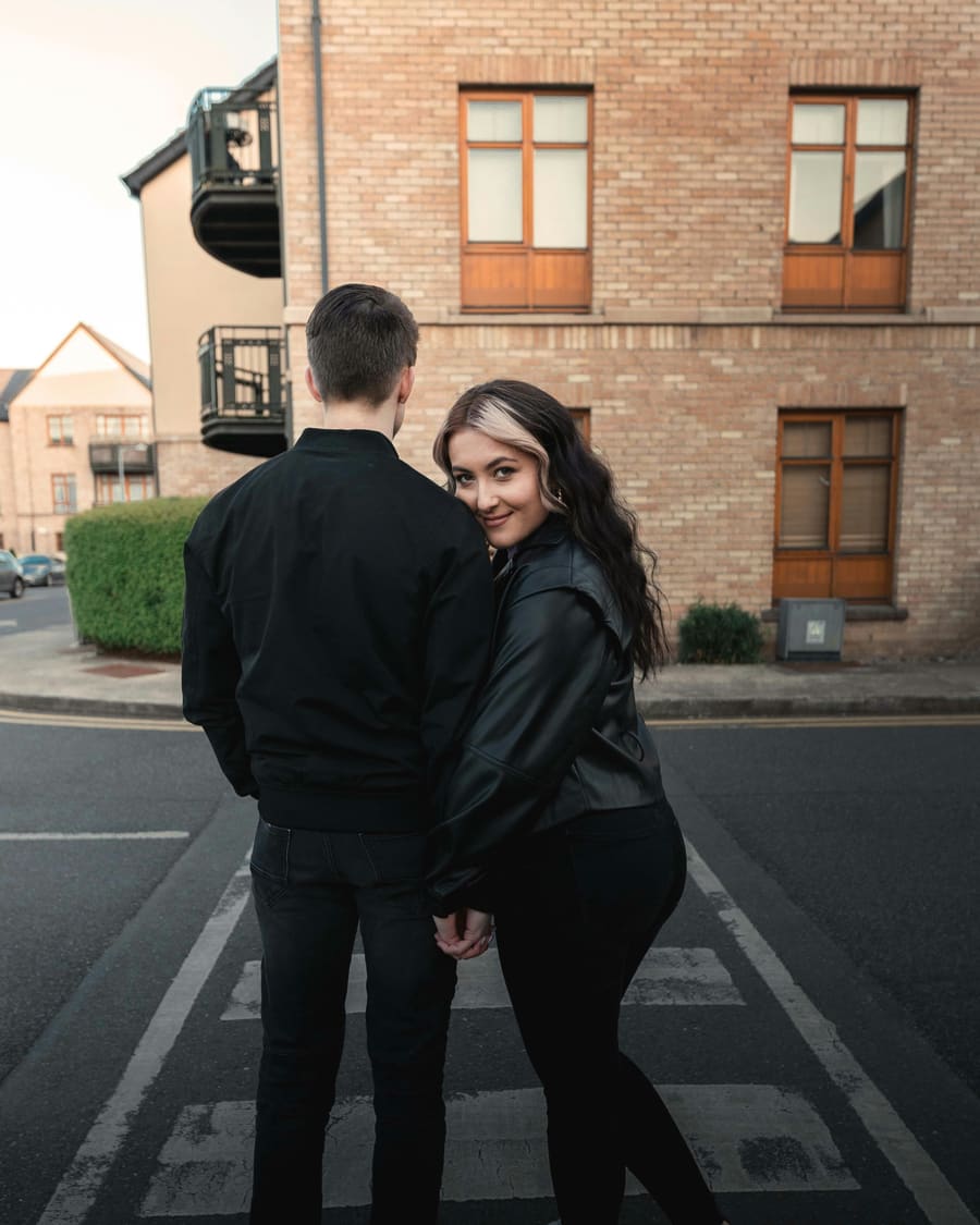A man in a black jacket walking a quiet street of brick houses with his date, a woman in a leather jacket who turns to smile at the camera.