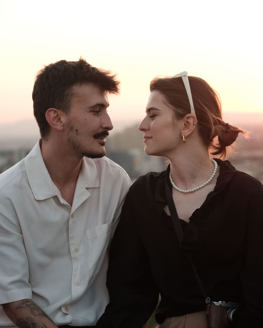 A man in a white shirt and his date, a woman wearing a pearl necklace, sharing a smile on a city rooftop as the sun sets.