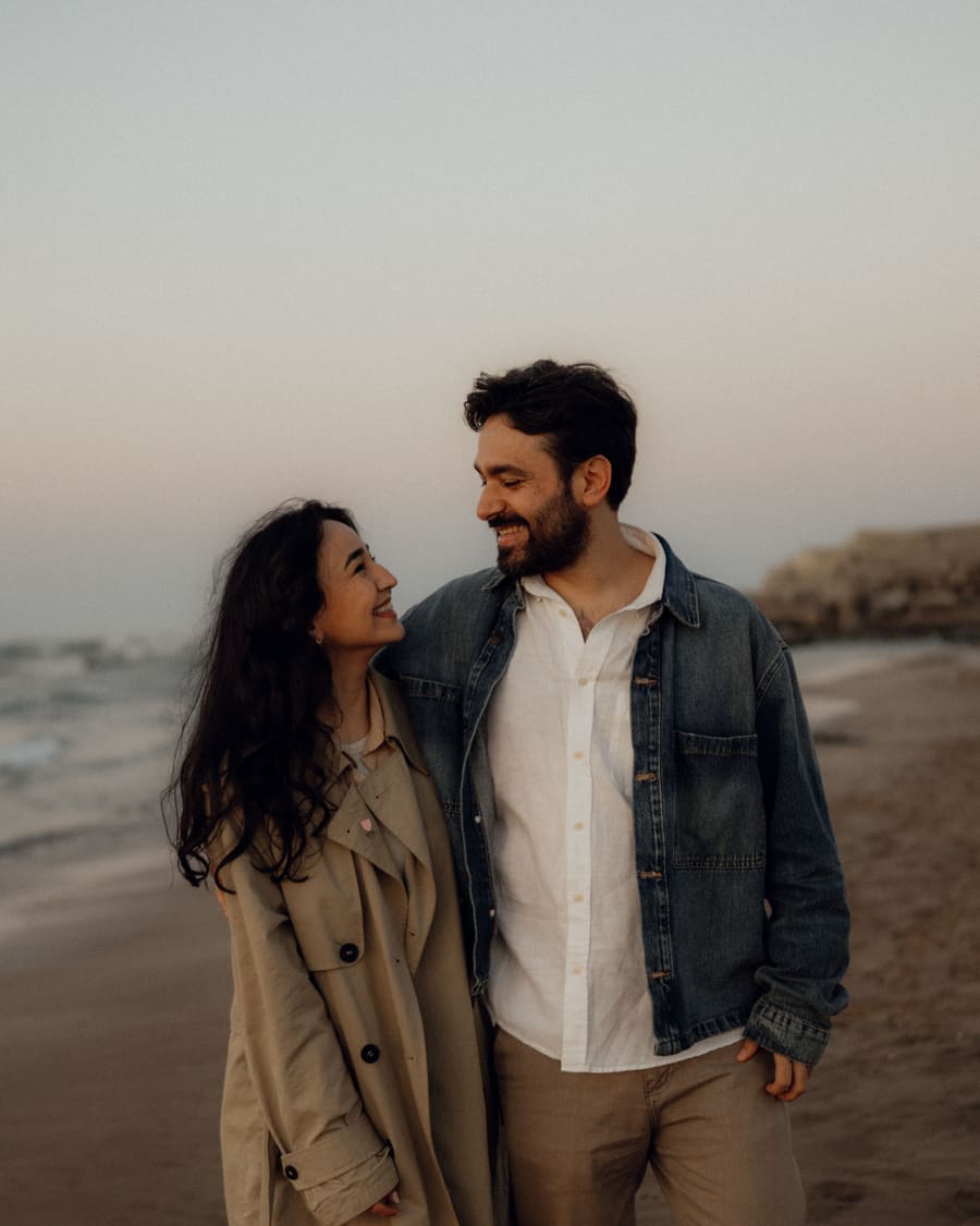 A man in a denim jacket and his date, a woman in a beige trench coat, laughing on the sand as dusk falls.