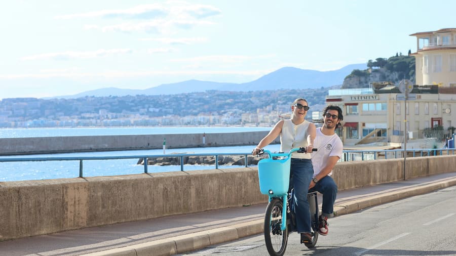 A man and his date, a woman in sunglasses, sharing one bicycle on a seafront promenade by a blue bay, both smiling.