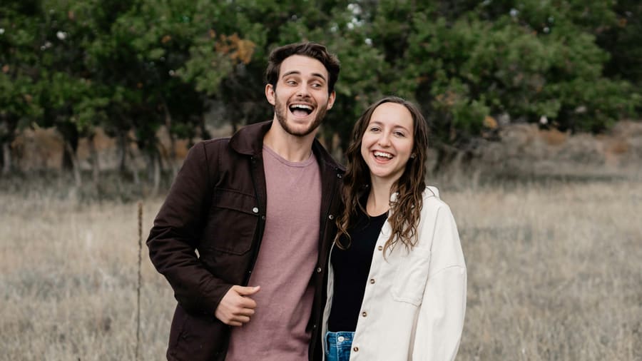 A man in a brown jacket and his date, a woman in a cream jacket, laughing side by side in a dry meadow with trees behind them.