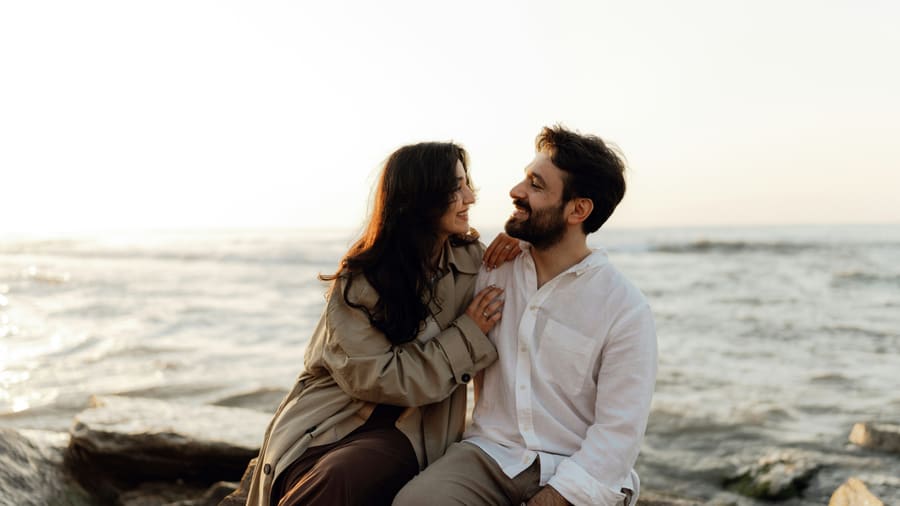 A man in a white shirt and his date, a woman in a trench coat, talking as they sit on seaside rocks in the low evening sun.