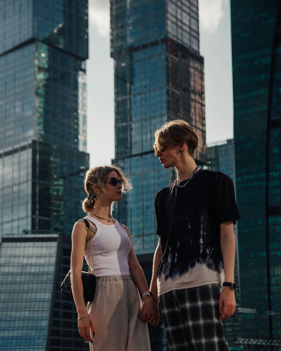 A man in a black T-shirt and his date, a woman in sunglasses and a white top, among tall glass office towers on a sunny day.